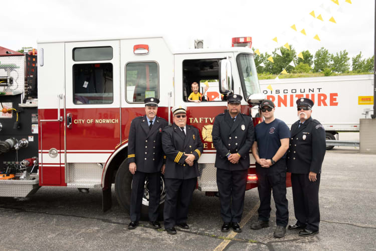 Five firefighters in uniform standing in front of a red and white City of Norwich fire truck with "TAFTVILLE FIRE" emblem visible.