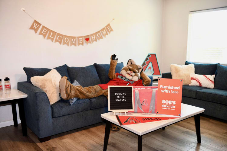 A living room with dark blue fabric sofas, a mascot in a red jersey lying on one sofa, a white marble coffee table with Arizona Diamondbacks merchandise, and signs reading "WELCOME TO THE DIAMOND" and "Furnished with love BOB'S DISCOUNT FURNITURE MYBOBS.COM."