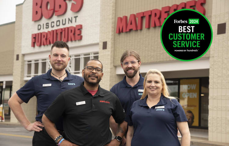 Four employees wearing Bob's Discount Furniture branded polo shirts stand outside a Bob's Discount Furniture store with red signage. A circular badge on the right reads "Forbes 2024 Best Customer Service Powered by hundredx."