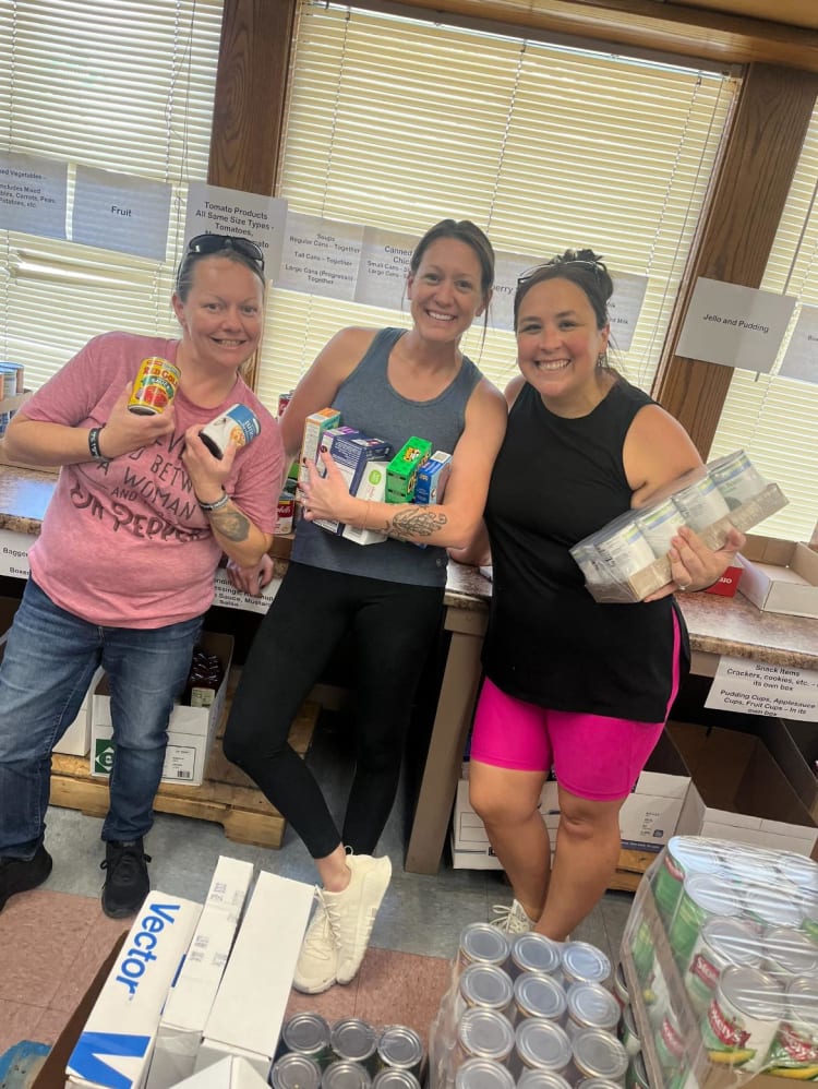 Three women standing indoors holding various canned and boxed food items, surrounded by labeled food pantry sections including Fruit, Tomato Products, Canned Chicken, Jello and Pudding, and Snack Items.