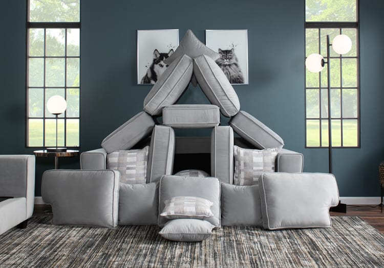 Stacked gray fabric sofa cushions arranged in a creative triangular shape in a modern living room with blue walls, large windows, and patterned rug.