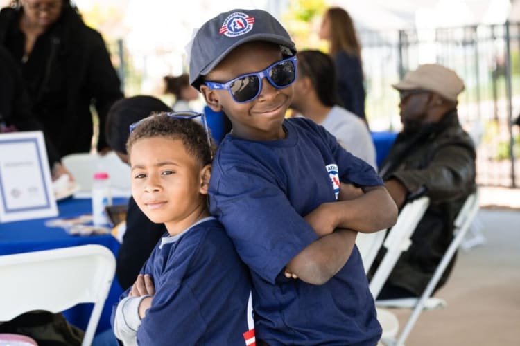Two boys wearing matching navy blue t-shirts and one with a navy blue cap stand back-to-back with arms crossed at an outdoor event with people seated at tables in the background.