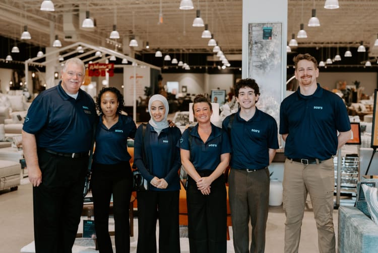Six retail employees wearing navy blue polo shirts with "BOB'S" logo stand inside a well-lit furniture store with sofas and decor in the background.