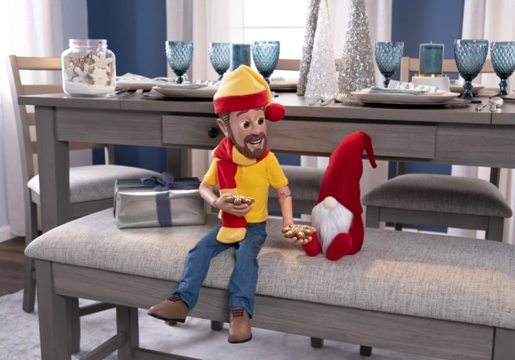 A festive holiday scene with a wooden doll wearing a yellow hat, red scarf, and blue jeans, holding decorated gingerbread cookies, seated on a gray cushioned bench next to a red gnome figurine with a white beard. The background features a gray dining table set with blue goblets, white plates, silverware, a silver wrapped gift, and decorative silver Christmas trees.