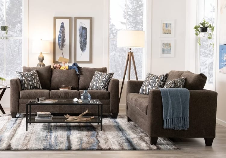 Modern living room setup with dark brown fabric sofas, geometric patterned cushions, glass-top black metal coffee table, tripod floor lamp, and feather wall art.