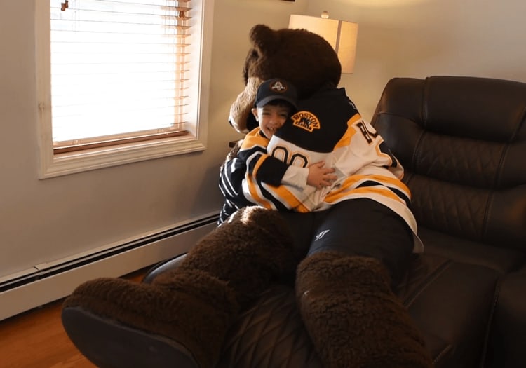 A person hugging a large brown bear mascot wearing a Boston Bruins hockey jersey, sitting on a black leather chair near a window with blinds.