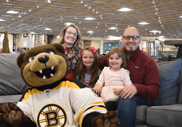 A person in a Boston Bruins mascot costume with a large brown bear head and white hockey jersey sits on a gray couch with three children and an adult wearing a red sweater in a spacious, well-lit indoor setting.