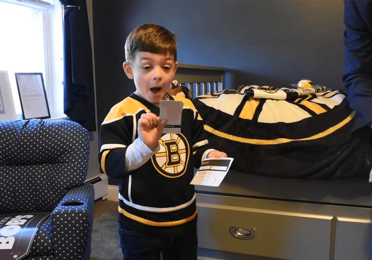 Young child wearing a black and yellow Boston Bruins hockey jersey holding two small cards, standing in a bedroom with matching Bruins bedding and decor.