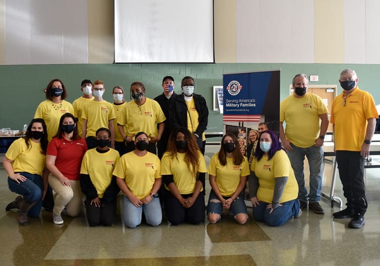 Group of volunteers wearing yellow and red shirts with "Ropp's Market" logo posing indoors in front of a banner that reads "Serving America's Military Families" with the Operation Homefront logo.