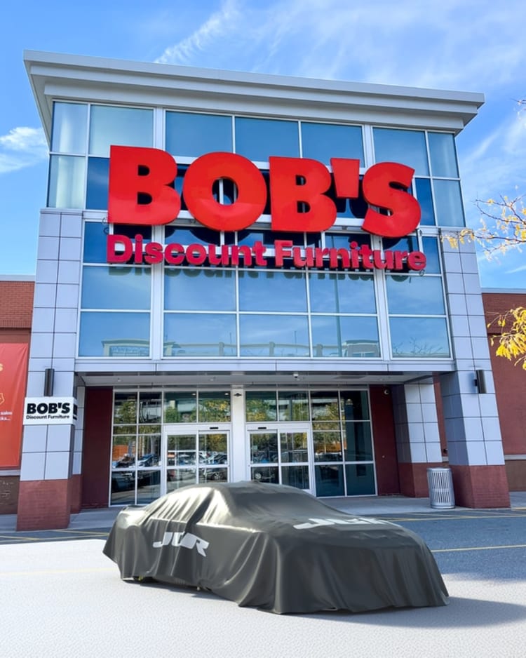 BOB'S Discount Furniture storefront with large red signage on glass facade and a car covered by a gray protective cover in front.