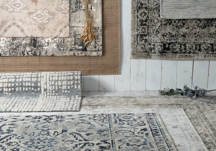 Layered rugs with various textures and patterns in neutral tones including beige, gray, and blue, displayed on a light wooden floor for home decor inspiration.