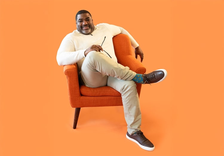 Man sitting on an orange upholstered armchair wearing beige pants, a cream sweater, blue patterned socks, and brown leather sneakers against an orange background.