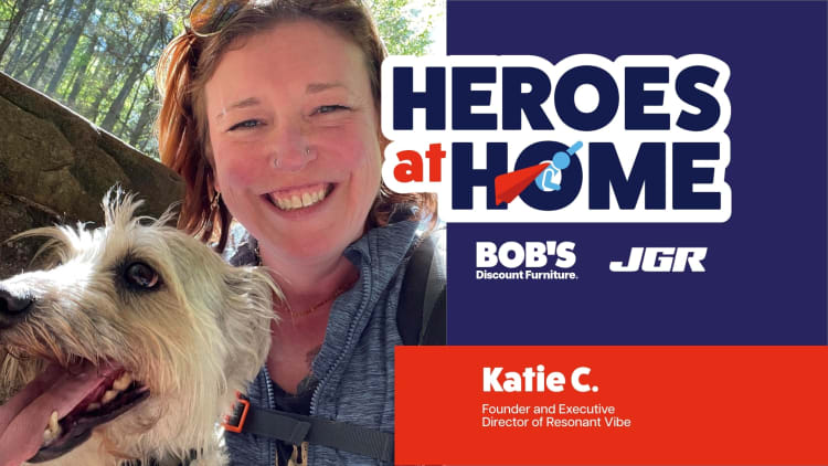 Woman with a light-colored dog outdoors next to a "Heroes at Home" banner featuring sponsors Bob's Discount Furniture and JGR, highlighting Katie C., Founder and Executive Director of Resonant Vibe.