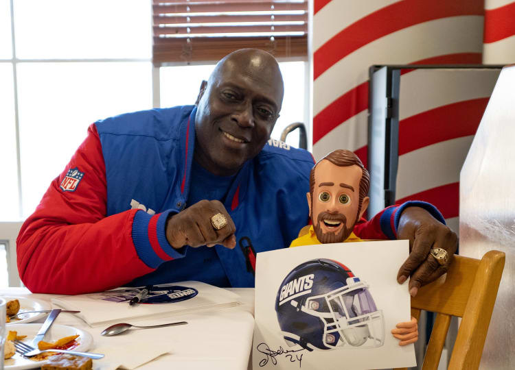 Man wearing a red and blue NFL jacket holding a signed print of a New York Giants football helmet with the signature "24" visible.