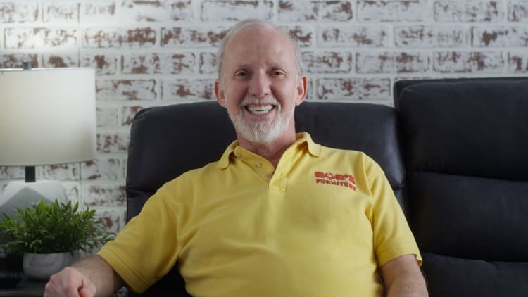 Man wearing a yellow polo shirt with "Bob's Furniture" logo in red, sitting on a dark gray sofa against a white brick wall.
