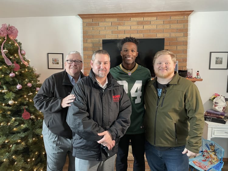 Four men standing indoors in front of a brick fireplace and TV, next to a decorated Christmas tree with pink ornaments and ribbon, wearing casual jackets and a Green Bay Packers jersey with number 74.