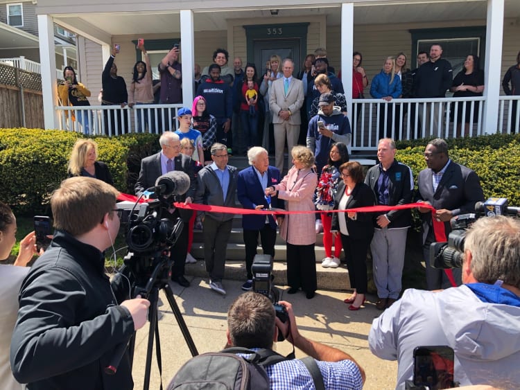 A ribbon-cutting ceremony in front of a house with a group of people, including officials and media, capturing the event.