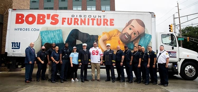 Group of uniformed emergency responders and staff standing in front of a large white truck with "BOB'S DISCOUNT FURNITURE" and "MYBOBS.COM" branding, featuring an advertisement of a person reclining on a blue chair.