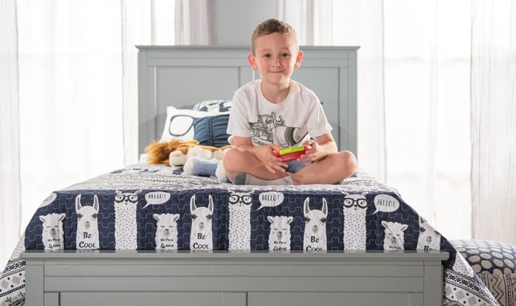 Child sitting cross-legged on a gray wooden bed with a navy blue and white llama-themed quilt featuring "Be Cool" and "Hello!" text, holding a handheld gaming device.