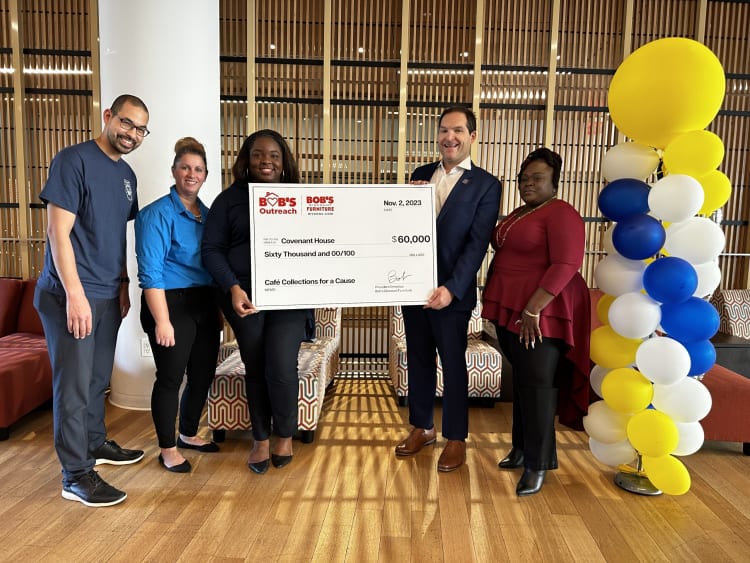 Five people standing indoors holding a large check from Bob's Outreach and Bob's Furniture, dated Nov. 2, 2023, for $60,000 payable to Covenant House, memo "Café Collections for a Cause," with yellow, white, and blue balloons on the right.