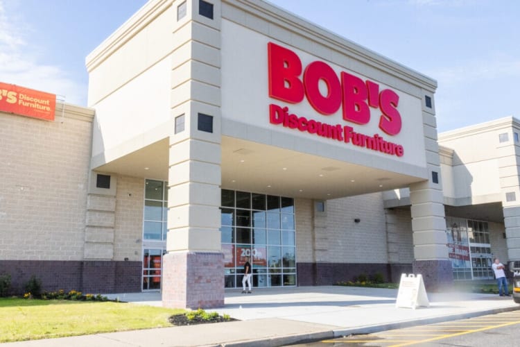 BOB'S Discount Furniture storefront with large red signage, beige and brick exterior, glass entrance doors, and a sidewalk ramp for accessibility.