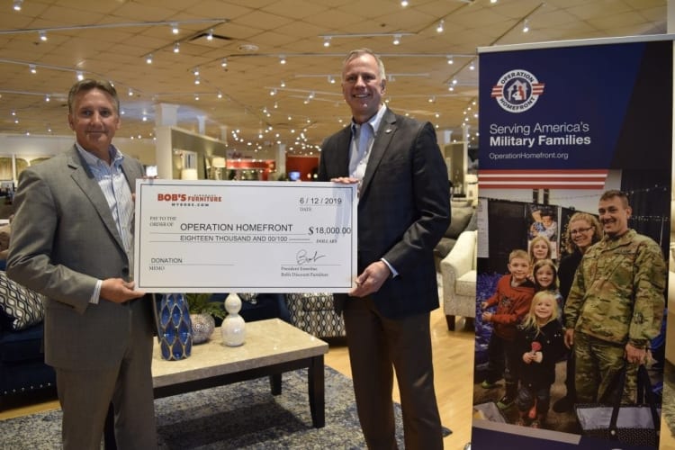 Two men in business attire hold a large check from Bob’s Furniture for $18,000 dated 6/12/2019, payable to Operation Homefront. Next to them is a banner with the Operation Homefront logo and text: "Serving America’s Military Families OperationHomefront.org," featuring an image of a military family. The setting appears to be a furniture showroom.