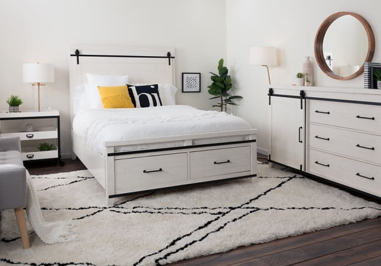 Modern bedroom with white wooden bed frame featuring black metal accents, white bedding, yellow and black decorative pillows, matching dresser with round mirror, and a plush white rug with black geometric patterns.