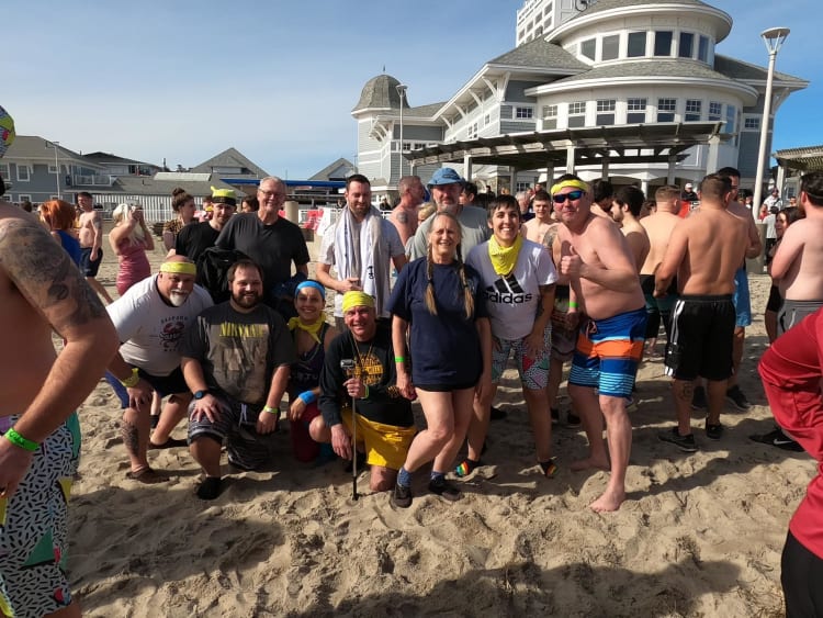 Group of people wearing casual beachwear and yellow bandanas gathered on sandy beach near a large white beach house under clear sky.