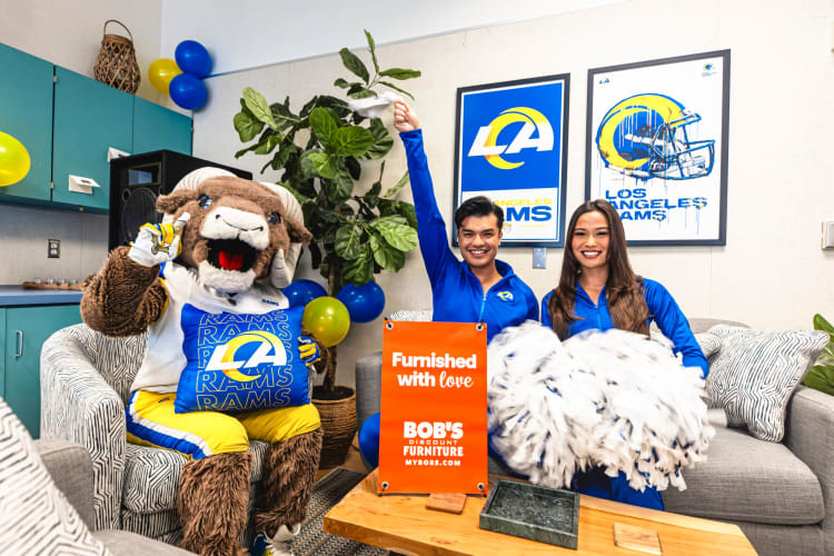 Los Angeles Rams mascot and two cheerleaders in blue uniforms holding pom-poms, sitting on a couch in a decorated room with Rams posters. An orange sign on the table reads: "Furnished with love BOB'S DISCOUNT FURNITURE MYBOBS.COM."