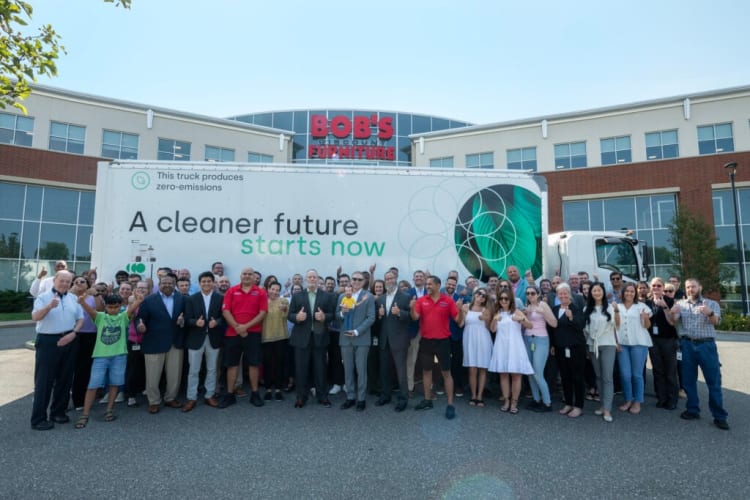 Group of diverse people standing in front of a large white truck with text "This truck produces zero-emissions. A cleaner future starts now" and Bob's Discount Furniture building in the background.