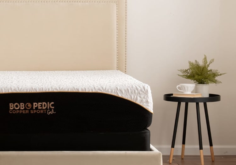 Mattress with beige upholstered headboard and black base, featuring "BOBOPEDIC Copper Sport Gel" branding on the side, next to a black round side table with wooden legs, a white cup, a book, and a green potted plant.