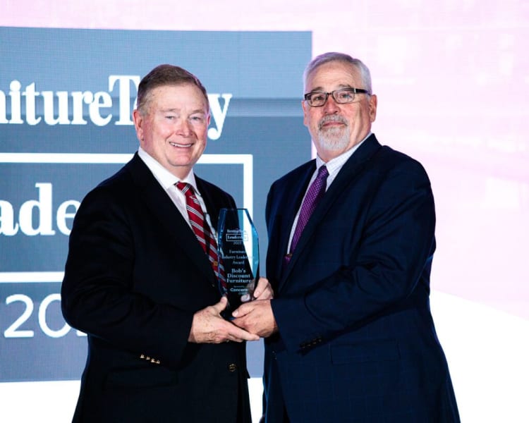 Two men in dark suits holding a clear glass award engraved with "Furniture Industry Leaders Award 2023, Bob's Discount Furniture, Concord, NH" in front of a backdrop with partial text "Furniture Today Leaders 2023."