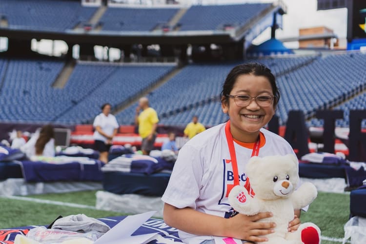 A person wearing a white t-shirt with a red lanyard sits in a stadium holding a white plush teddy bear with red paw prints and feet.