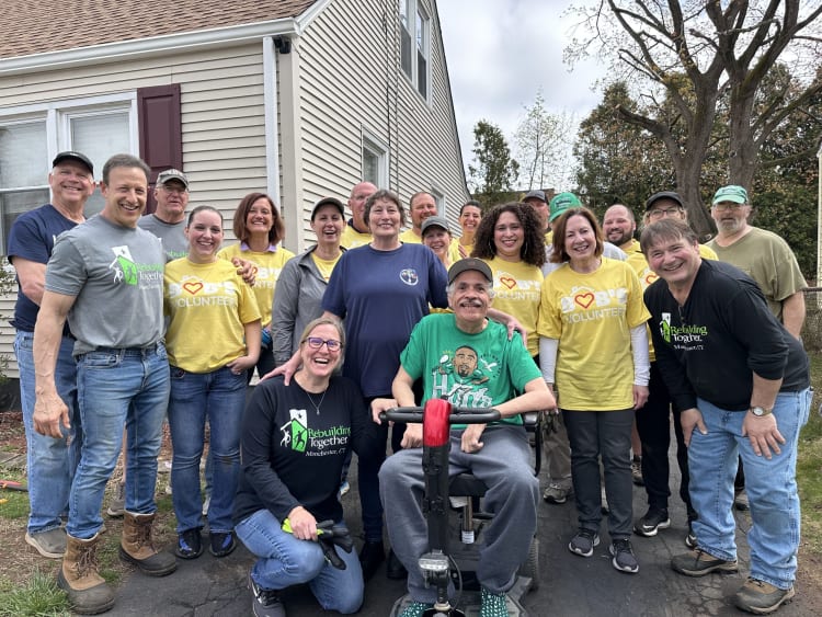 Group of volunteers wearing "Rebuilding Together Manchester, CT" and "Big Hearts Volunteer" shirts posing outdoors in front of a house, showing community teamwork.