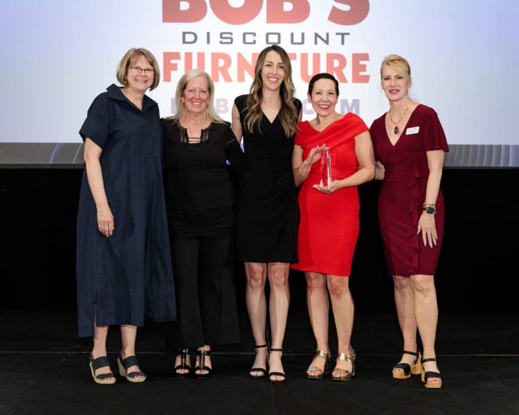 Five women stand on stage in front of a "Bob's Discount Furniture" logo backdrop. They wear formal attire: navy dress, black outfit, black dress, red dress holding an award, and burgundy dress. The setting suggests an award ceremony or corporate event.