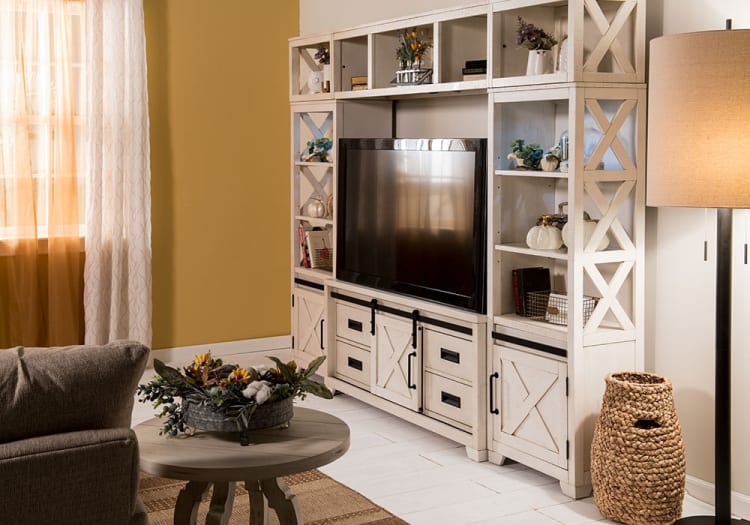 White wooden entertainment center with open shelves and cabinets, housing a flat-screen TV, decorative plants, and storage baskets, next to a woven basket and floor lamp in a cozy living room with beige walls and sheer curtains.