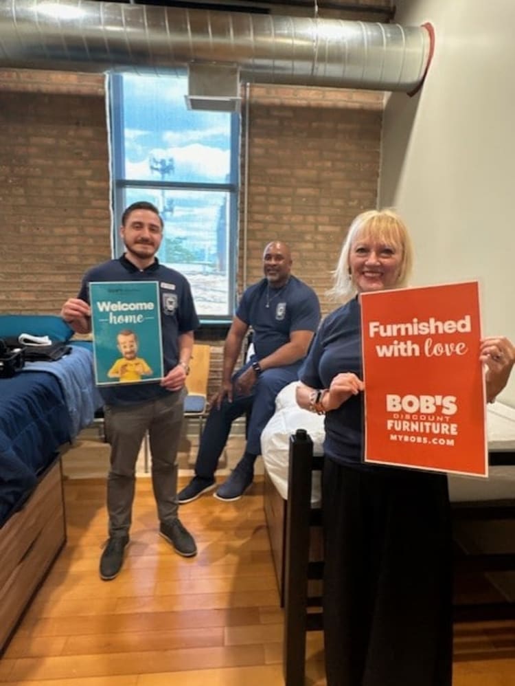 Three people in a dorm room holding signs; one reads "Welcome home" with a cartoon character, the other "Furnished with love BOB'S DISCOUNT FURNITURE MYBOBS.COM," promoting furnished living spaces and Bob's Discount Furniture.