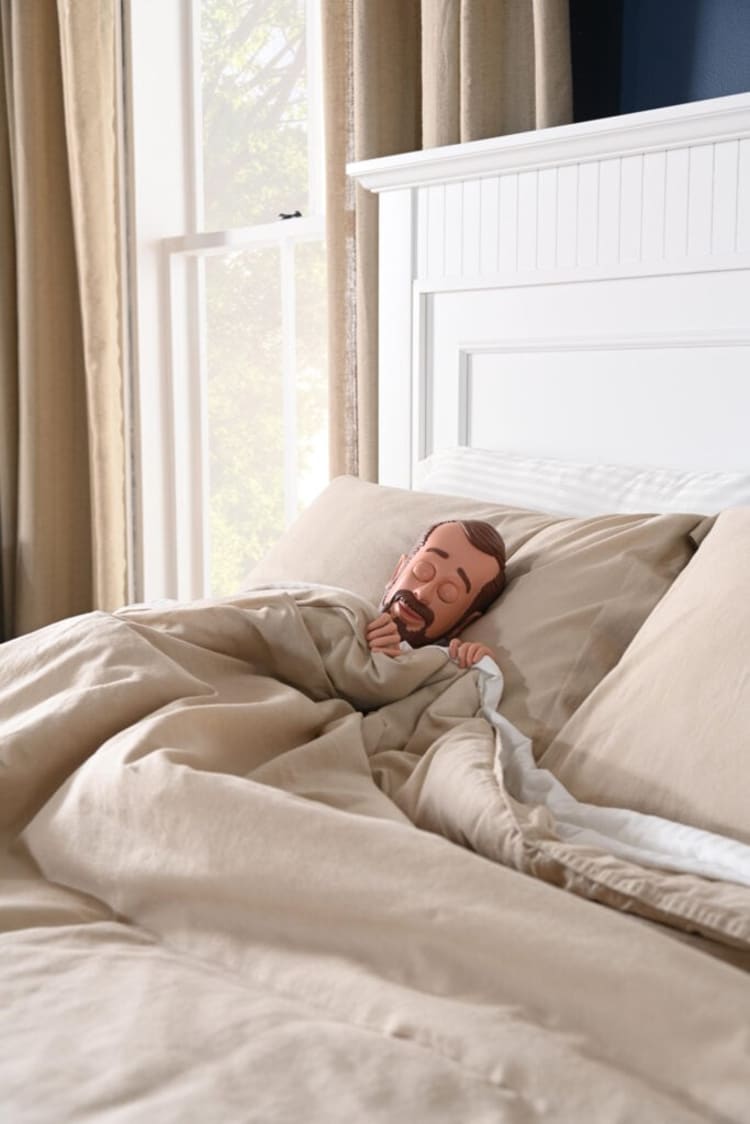 Child tucked in a beige linen bed with matching pillows and a white wooden headboard, next to a window with beige curtains.