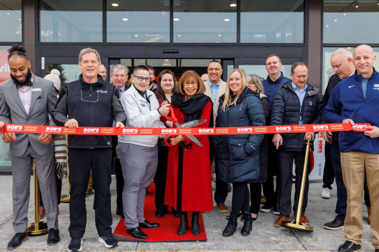 Group of people at a grand opening ribbon-cutting ceremony outside Bob's Discount Furniture, holding a red ribbon with "BOB'S GRAND OPENING!" text.