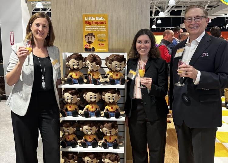 Three people holding drinks stand beside a display of plush toys called Little Bob, featuring a cartoon character with brown hair, yellow shirt, and blue pants. A sign above reads: "Little Bob, Big Impact. Take Little Bob home and help give shelter to those in need in our community." The setting appears to be an event or promotional space with bright lighting and a modern interior.