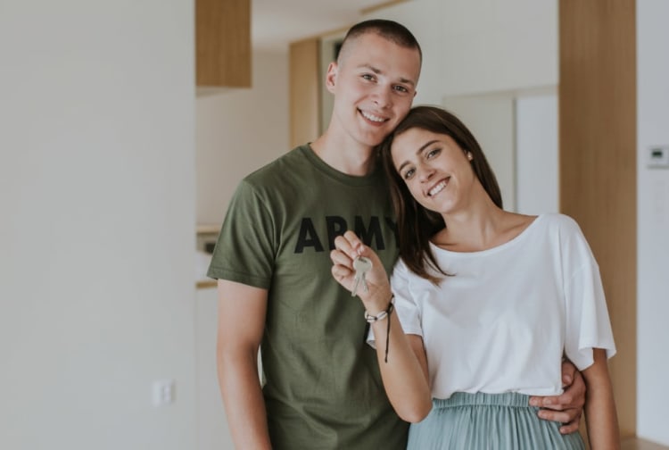 A couple standing indoors, the woman holding a key, symbolizing homeownership or moving into a new house. The man wears an olive green shirt with "ARMY" text, and the woman wears a white top and light blue skirt.
