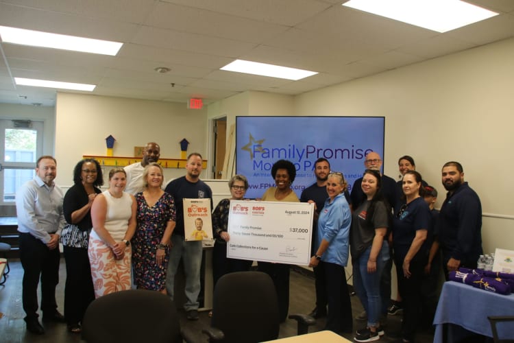 A group of people standing indoors holding a large check for $37,000 dated August 12, 2024, from Bob's Outreach to Family Promise, with a TV screen in the background displaying "Family Promise" and related text.