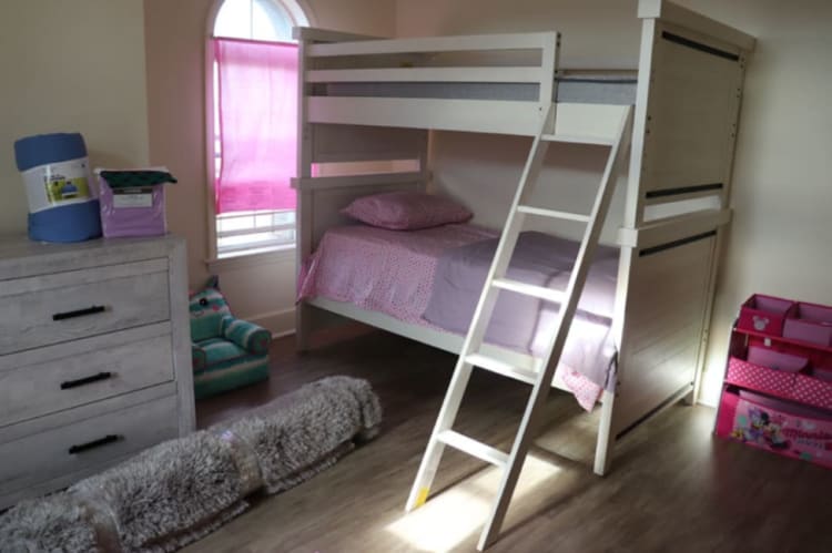 White wooden bunk bed with pink and purple bedding in a child's bedroom, featuring a ladder, dresser, and Minnie Mouse-themed storage organizer.