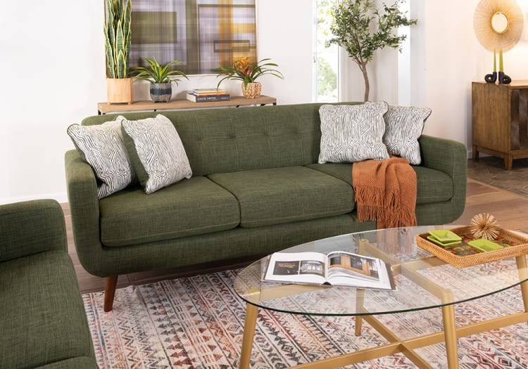 Green mid-century modern fabric sofa with wooden legs, patterned white throw pillows, orange throw blanket, glass coffee table, and decorative plants.