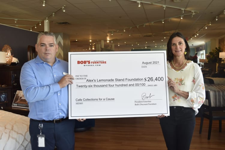 Two people holding a large check from Bob's Discount Furniture dated August 2021, paying $26,400 to Alex's Lemonade Stand Foundation for Cafe Collections for a Cause.