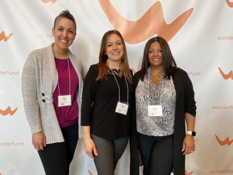 Three women standing in front of a Wonderfund branded backdrop, wearing business casual attire with name badges, posing for a group photo.