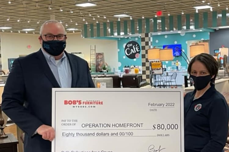 Two people holding a large check from Bob's Furniture dated February 2022, payable to Operation Homefront for $80,000, with the amount written as "Eighty thousand dollars and 00/100." The background shows a café area with tables and chairs.