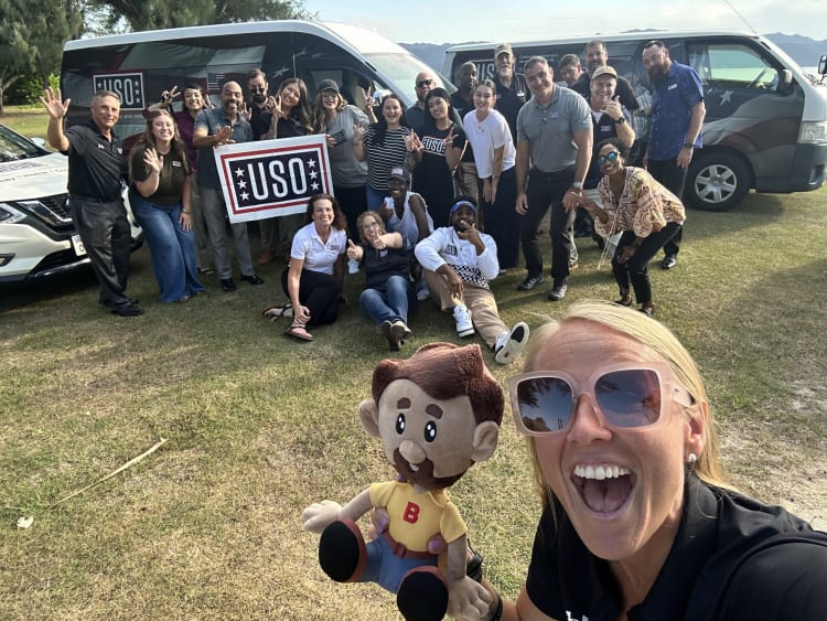Group of people outdoors holding a USO sign near vans with USO logos, with one person holding a plush doll with brown hair, beard, yellow shirt, and blue pants.