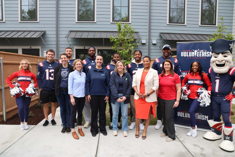 Group of people posing outdoors in front of a gray building with windows, including individuals in New England Patriots jerseys, cheerleader outfits, and casual attire, standing near a banner that reads "Celebrate Volunteerism Patriots Foundation."