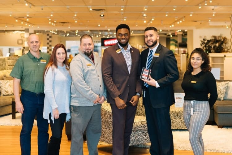 Six retail employees standing in a furniture store showroom, dressed in business casual and formal attire, holding an award plaque.